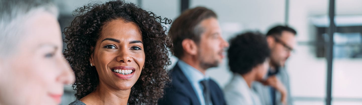 A beautiful and smiling woman is seated at a boardroom table surrounded by fellow board members. 