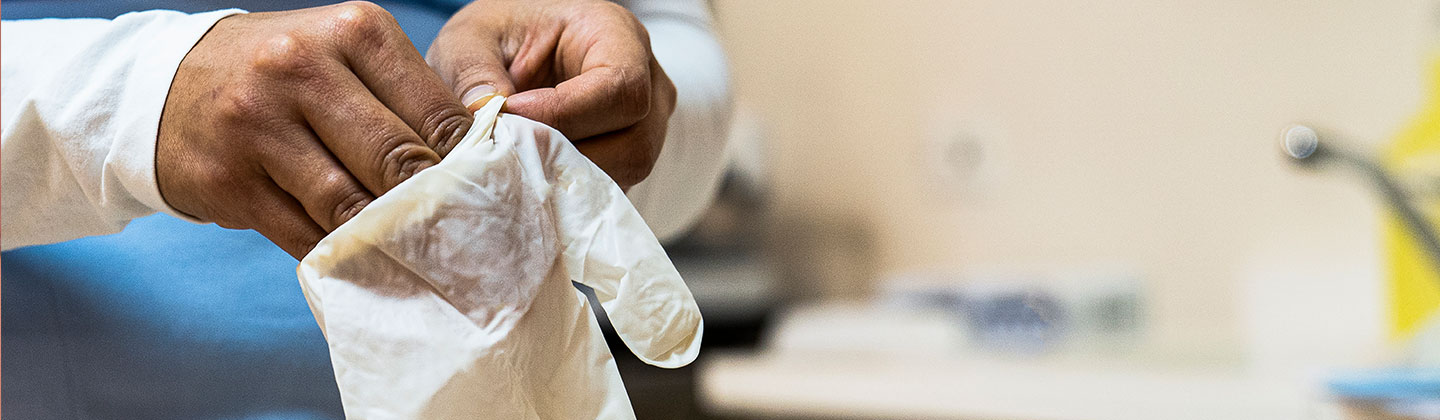 A Nurse wearing blue scrubs puts her their hand into a exam glove. Only their body, arm and hand are visible. 