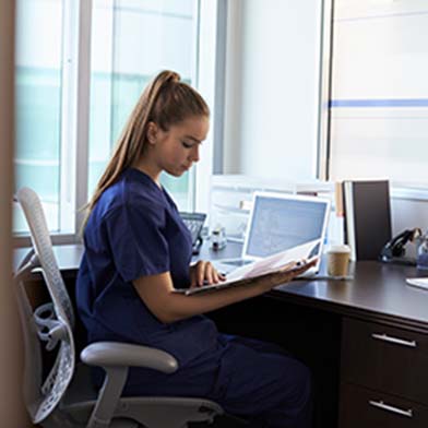 A%20female%20nurse%20wearing%20dark%20blue%20scrubs%20is%20seated%20at%20a%20desk%20and%20reading%20information%20from%20a%20folder.%20A%20computer%20screen%20can%20be%20seen%20in%20front%20of%20her.