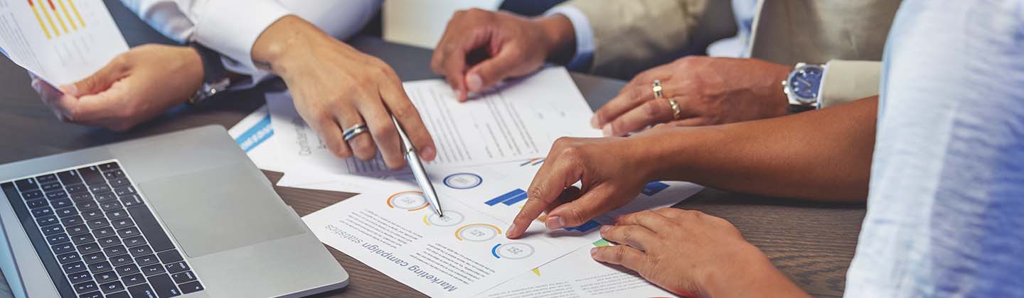 Three people are seated at a table and reviewing financial data spread out before them. Only their arms and hands are visible as they point to sections of a graphic. 