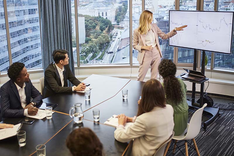 A female nurse manager is standing at the front of a conference room, and pointing to trend data on a white board which is set up behind her. Other people are seated at a conference table and listening to the presentation.