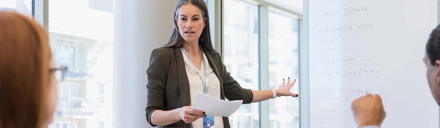 A female Director of Nursing stands in front of a conference table and is looking at a group of seated employees while giving a presentation and pointing to some information on a whiteboard behind her.