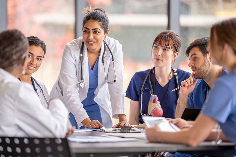 A group of attentive nurses in scrubs and white coats gather around a table for a team briefing, led by a nurse standing up, indicating a collaborative medical discussion.