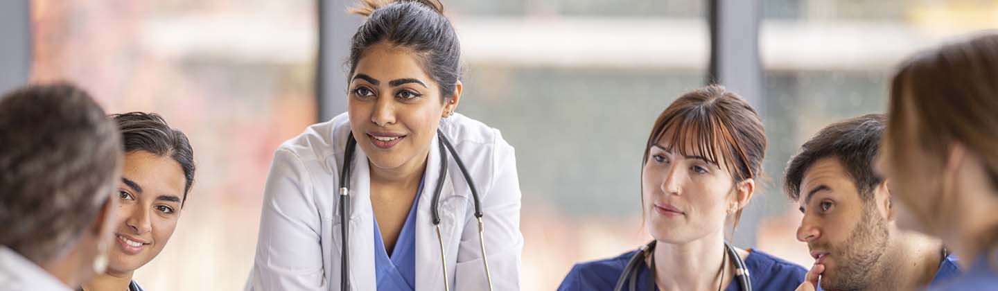 A group of attentive nurses in scrubs and white coats gather around a table for a team briefing, led by a nurse standing up, indicating a collaborative medical discussion.