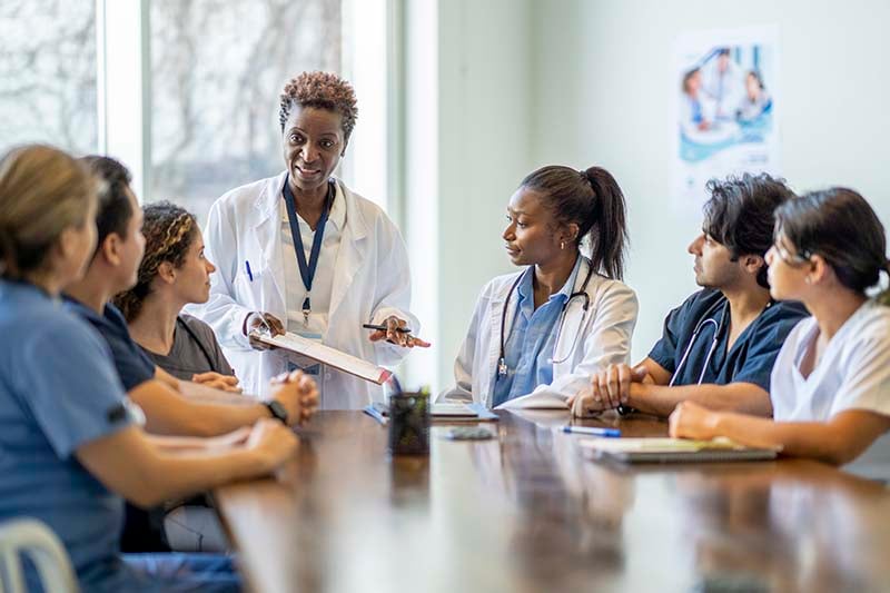 A diverse team of nurses gathers around a table for a meeting, with one standing and leading the discussion, illustrating collaboration and engagement in a clinical setting.