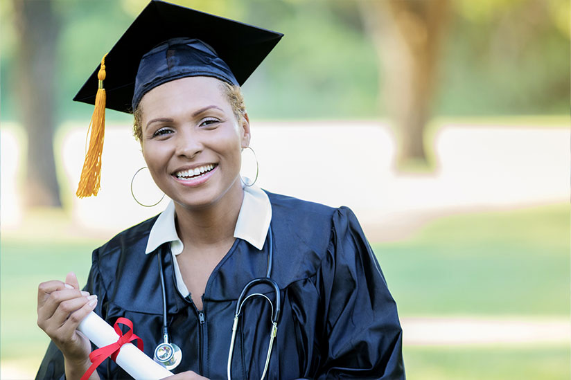 Smiling young nurse with an afro holds a clipboard in a classroom with fellow nurses chatting behind.