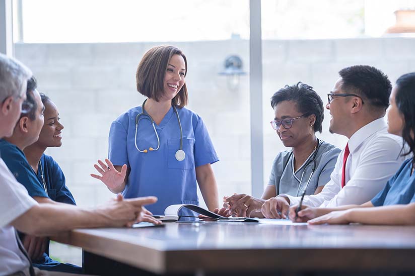 A young female nurse is wearing blue scrubs and is standing at the head of a conference table in front of a group of seated health care workers. She is smiling and appears to be sharing a story.