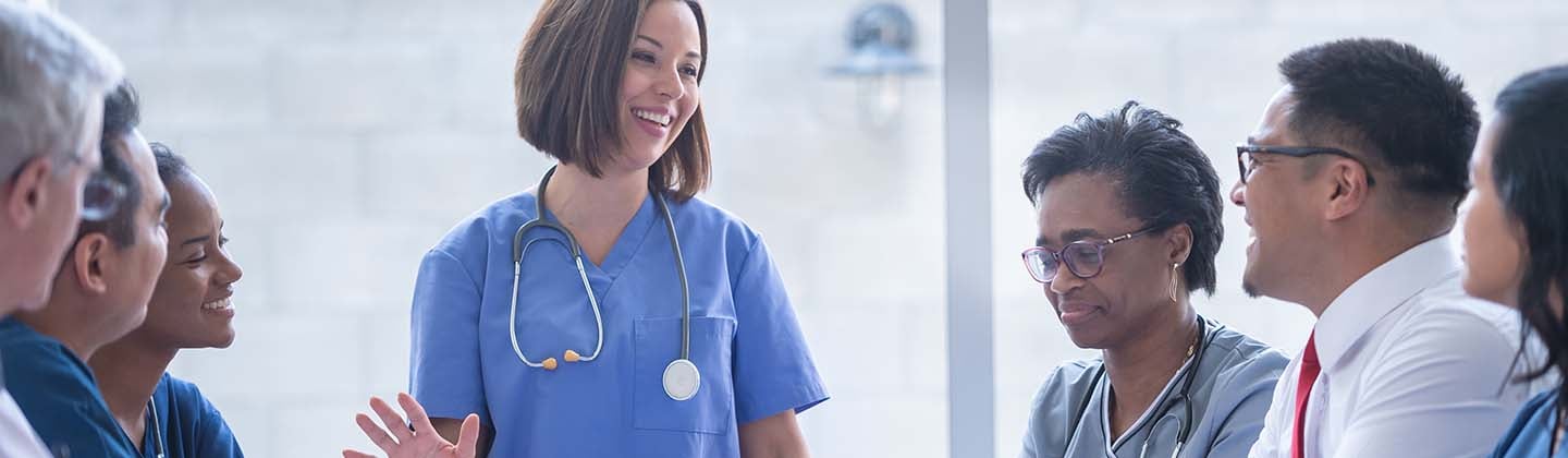 A young female nurse is wearing blue scrubs and is standing at the head of a conference table in front of a group of seated health care workers. She is smiling and appears to be sharing a story.