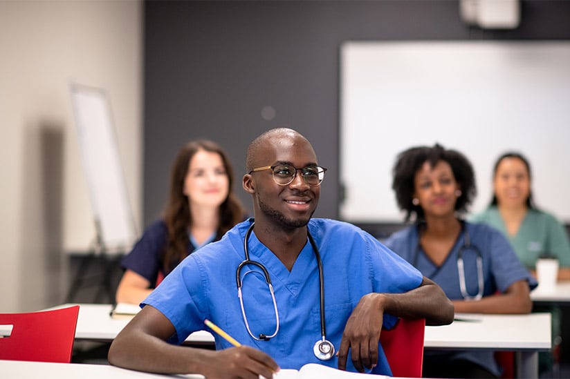 A diverse group of nursing students dressed in various color scrubs are engaged in learning in a classroom setting.