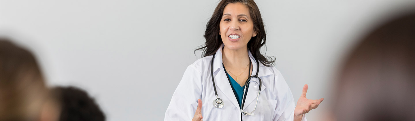 A nurse educator in mid-lecture, standing confidently in front of a classroom. She is dressed in a white lab coat with a stethoscope around her neck, gesturing openly to her students. Her expression is enthusiastic and engaging, suggesting an interactive teaching moment. The blurred foreground shows the back of students’ heads, focusing attention on the educator.