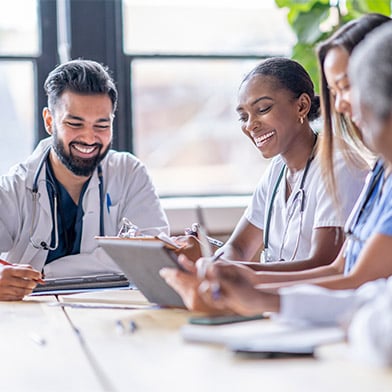 A small group of four medical professionals sit around a boardroom table as they meet to discuss patient cases. They are each dressed professionally in scrubs and lab coats as they focus on working together.