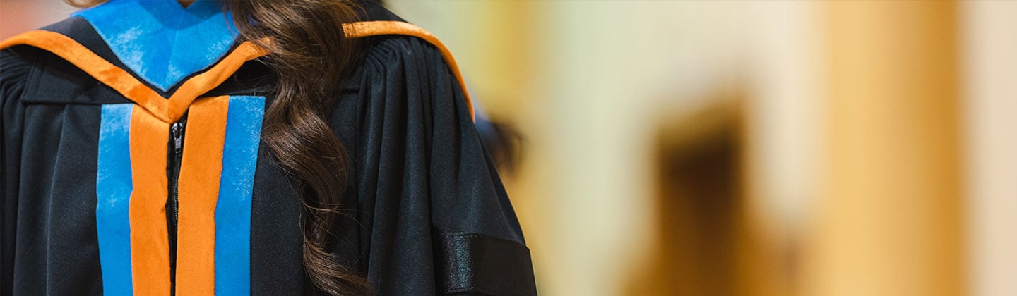  Image of a doctoral student standing while wearing their black graduation gown which has an orange and blue hood and front detail. Only their body is visible. 