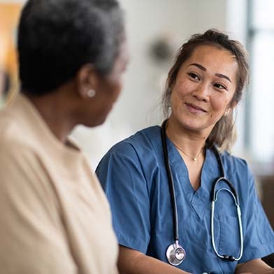 A cheerful nurse in blue scrubs takes notes on a clipboard while engaging with an elderly patient in a clinic room, signifying a caring and professional nurse-patient interaction.