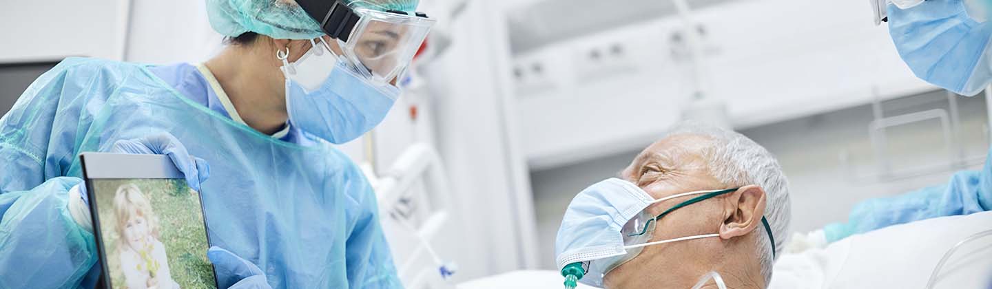 Two nurses are wearing isolation gowns, goggles, and facemasks, and are standing at either side of a patient’s bed. An elderly male patient is in the bed. He is also wearing a mask, and he is looking at a tablet screen the nurse is holding so he can talk with his family over the internet.