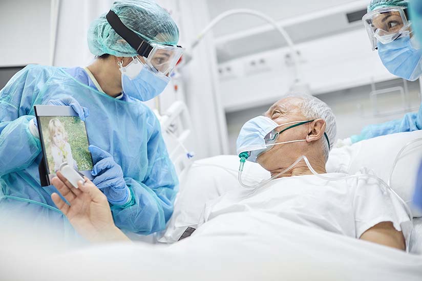 Two nurses are wearing isolation gowns, goggles, and facemasks, and are standing at either side of a patient’s bed. An elderly male patient is in the bed. He is also wearing a mask, and he is looking at a tablet screen the nurse is holding so he can talk with his family over the internet.