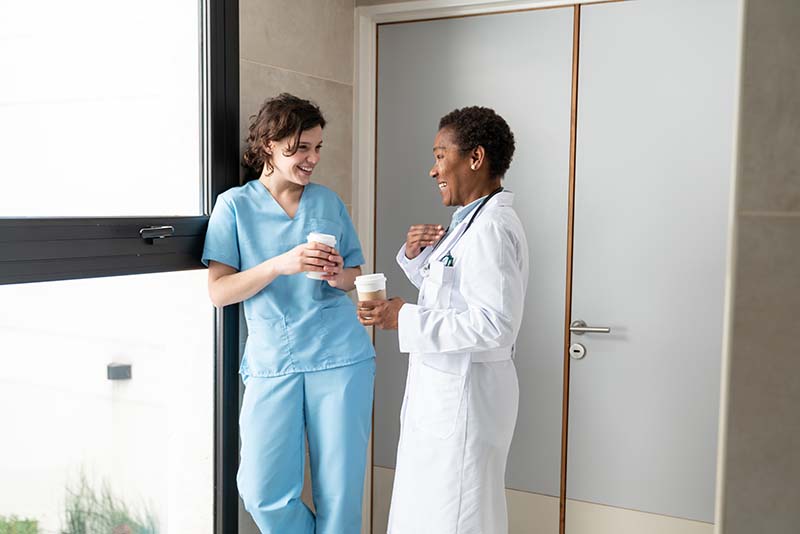Two nurses are standing in front of a doorway and are deep in conversation. One is wearing a white lab coat, and the other is dressed in scrubs, and they are both drinking from white cups. They are relaxed and smiling.