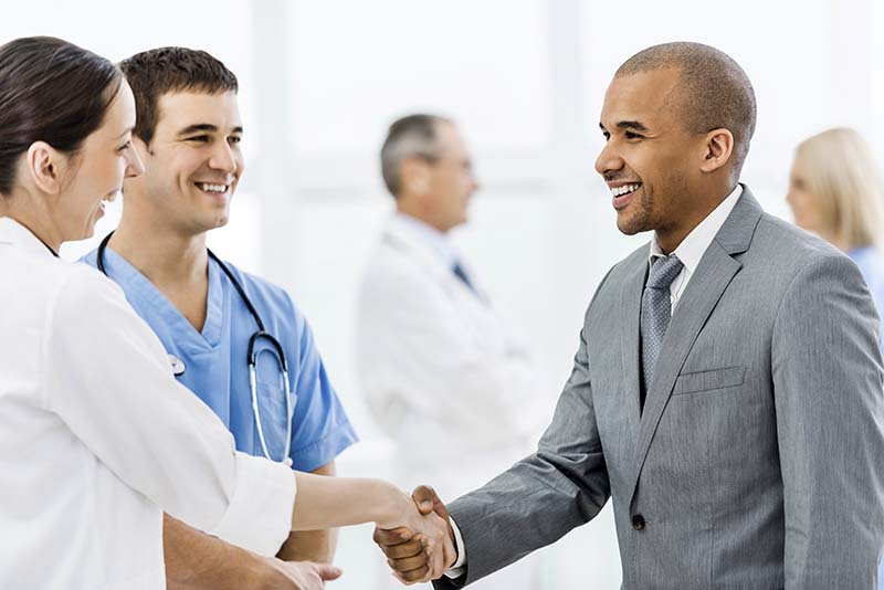 Two nurses in scrubs, one male and one female, are smiling and welcoming a man in a gray suit with a handshake. In the background, another healthcare professional is visible. The interaction occurs in a bright, clinical setting, suggesting a positive professional encounter.