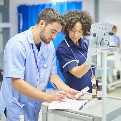 Two nurses are standing at a medication cart in the Recovery Room. A male nurse is holding a box of medication and doing documentation in a logbook. A female nurse is verifying the correct medication and entering information on a computer screen.