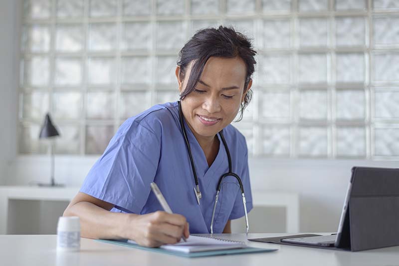 A young female nurse is seated at a desk, wearing a light blue scrub outfit and doing research using a laptop and taking notes.