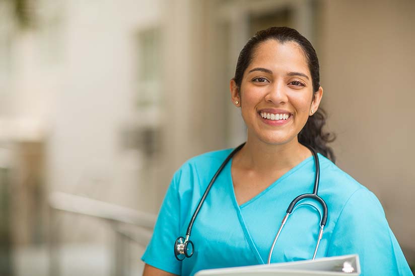 A cheerful nurse in turquoise scrubs, with a stethoscope around her neck, holds a clipboard and smiles warmly, standing in a well-lit hospital corridor.