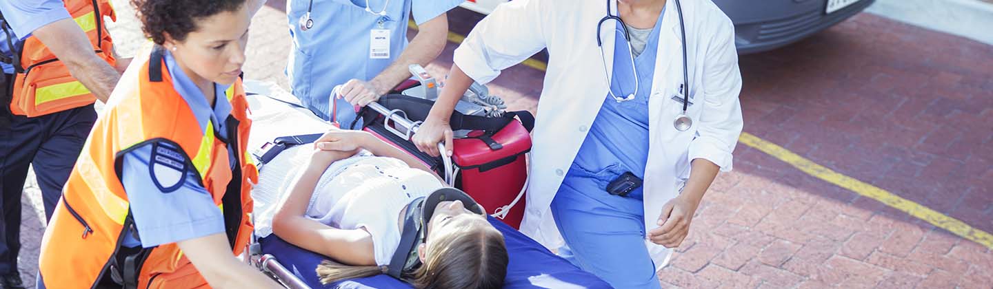 Two Emergency Room staff members are helping a team of emergency medical technicians to push a stretcher into the Emergency Department. A young child wearing a neck brace is calmly laying on the stretcher.