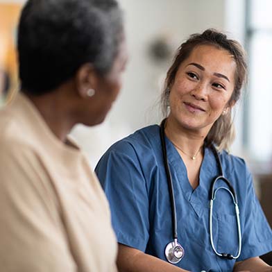 A cheerful nurse in blue scrubs takes notes on a clipboard while engaging with an elderly patient in a clinic room, signifying a caring and professional nurse-patient interaction.