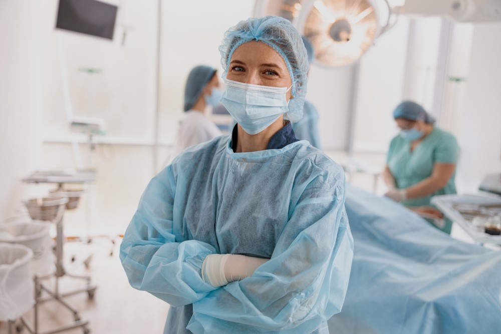 A nurse is dressed in a surgical gown, hair covering, surgical gloves and mask. She is standing with arms folded in the middle of an operating room suite. A patient and another nurse can be seen in the background.
